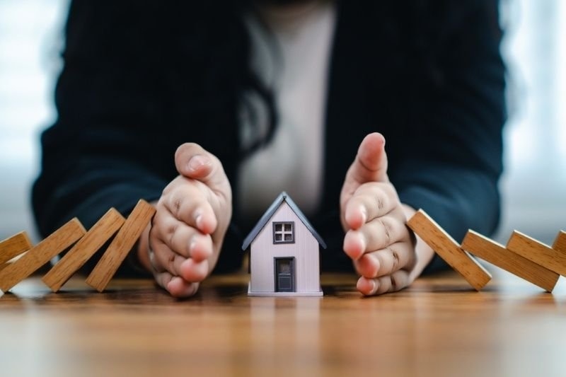 Hands shielding a miniature house from falling dominoes.