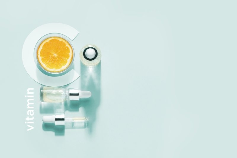 Flat-lay arrangement featuring an orange slice, cosmetic bottles, and droppers with “vitamin” text on a light background.
