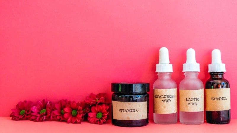 Skincare products labeled Vitamin C, Hyaluronic Acid, Lactic Acid, and Retinol arranged with red flowers against a bright pink background.