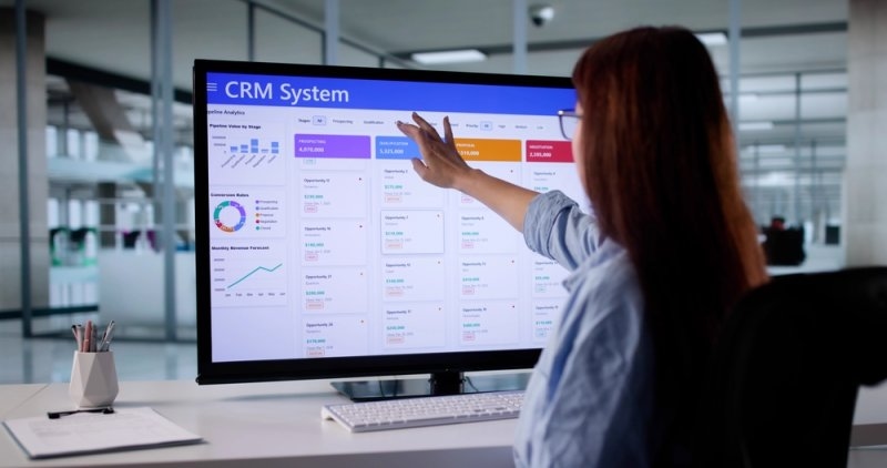 Professional analyzing a CRM system dashboard with sales pipeline stages, conversion rates, and revenue forecasting charts on a large monitor.