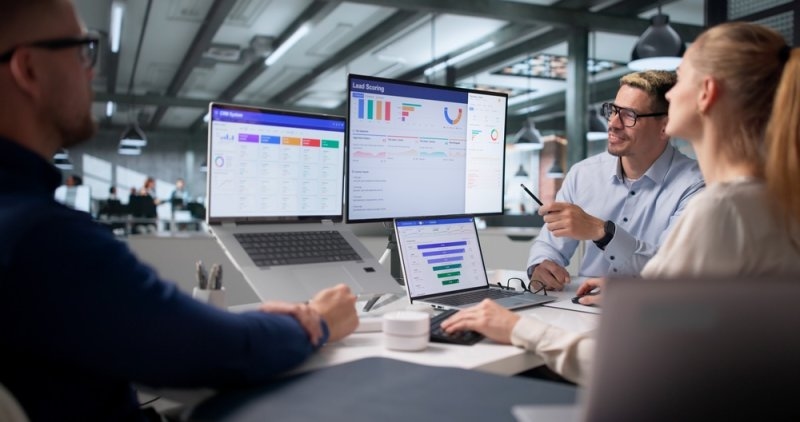 Business team collaborating around multiple screens displaying CRM dashboards, lead scoring analytics, and sales performance data.