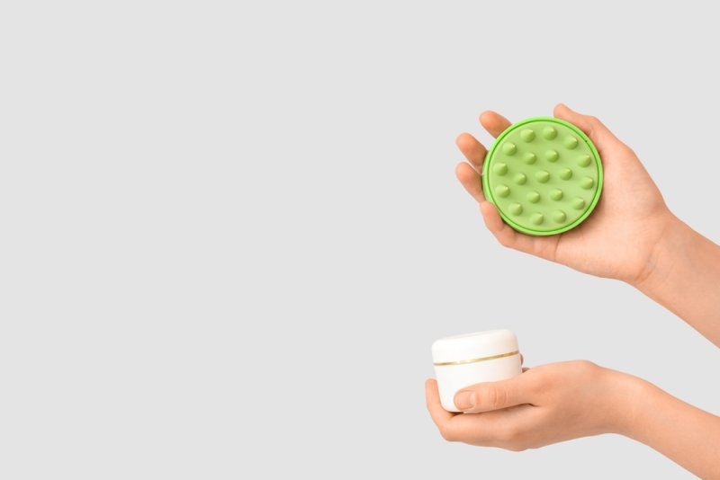 Green silicone scalp massager and a white hair-care product jar held against a light grey background.