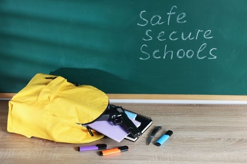 Classroom scene with a backpack, school supplies, and a handgun on a desk beneath the words “Safe Secure Schools” written on a chalkboard.