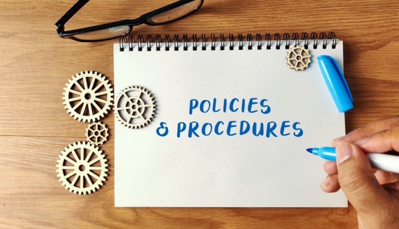 Hand writing Policies and Procedures on a notebook with wooden gears and glasses on a wooden desk symbolizing rules and systems.