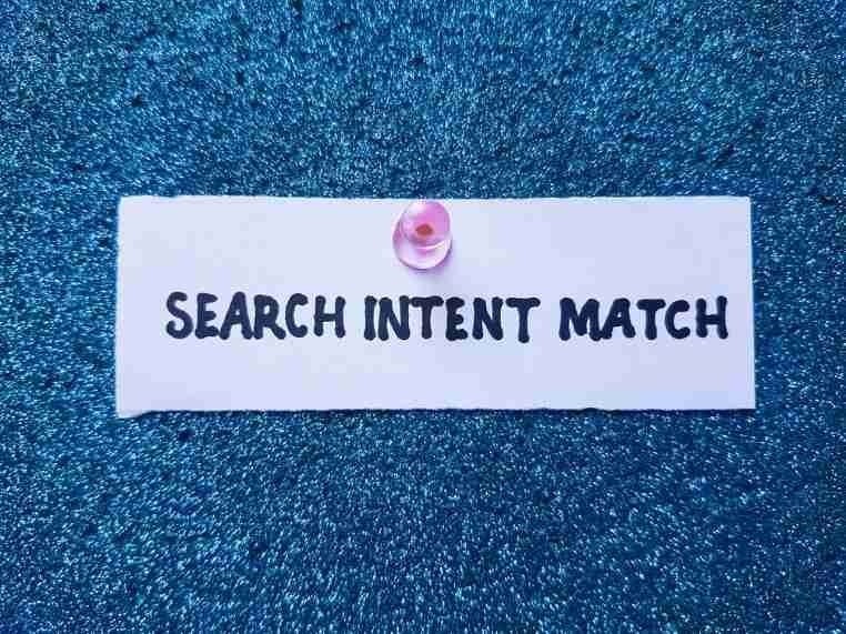 A small white card pinned to a blue textured surface displays the words “Search Intent Match.”