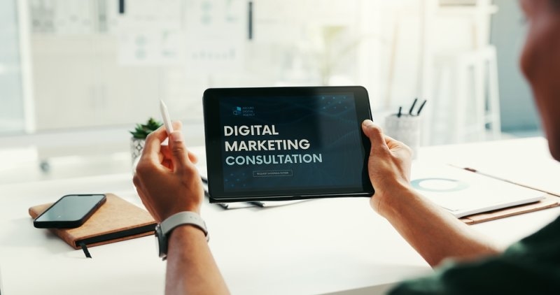 Person holding a tablet displaying “Digital Marketing Consultation” at a desk with a smartphone and business materials.