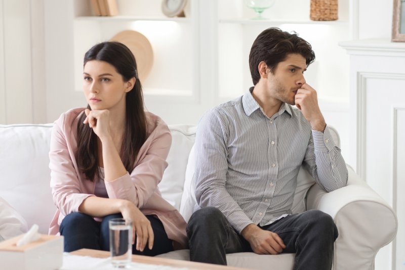 A young couple sits on a sofa in a brightly lit living room. Both appear to be lost in thought, staring off into the distance with serious expressions. 