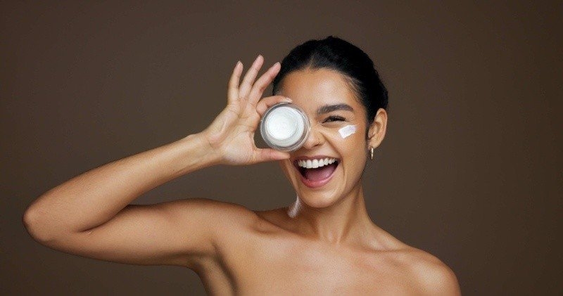 Smiling woman holding a skincare cream jar over one eye while applying moisturizer to her cheek.