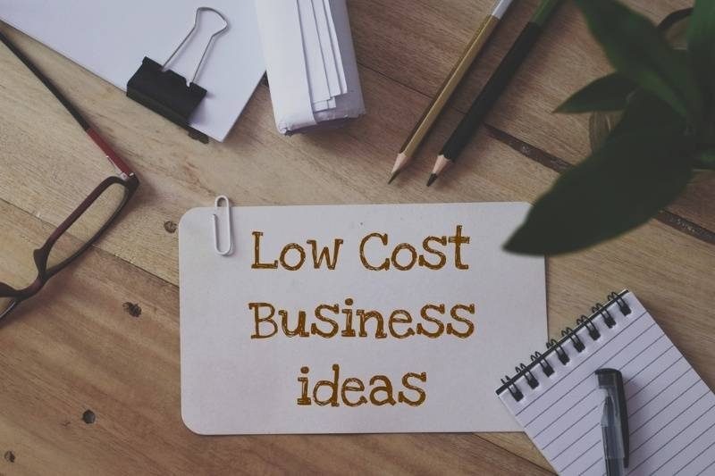 A paper card reading “Low Cost Business Ideas” sits on a wooden desk surrounded by pens, notebooks, paper clips, and office supplies.