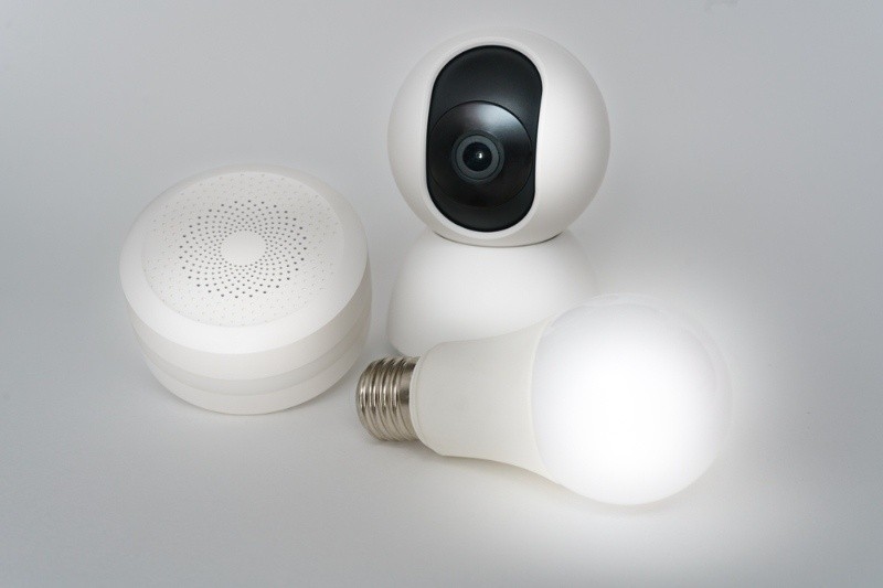 Smart home devices including a security camera, smart speaker, and smart light bulb on a white background.