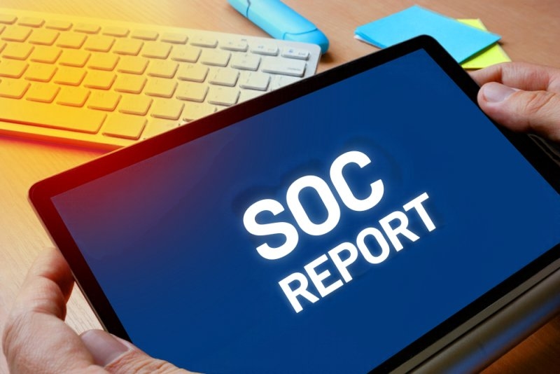 Hands holding a tablet displaying “SOC Report” on a desk.