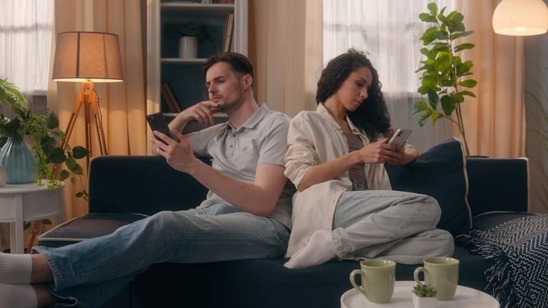 Couple sitting back-to-back on a couch, both using smartphones, suggesting disconnection or lack of communication.