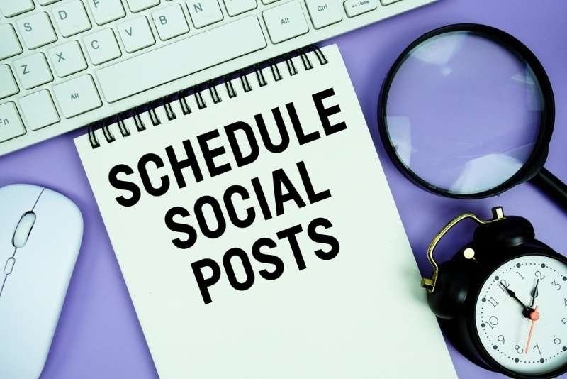 Notepad with the text "Schedule Social Posts" and white keyboard, wireless mouse, magnifying glass and alarm clock.