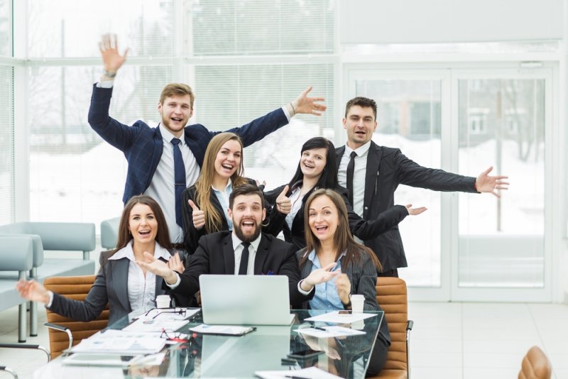 Diverse group of business professionals smiling and celebrating together around a laptop in an office.