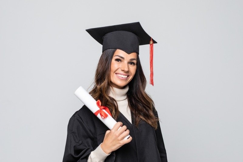A smiling young woman wearing a black graduation cap and gown holds a rolled diploma tied with a red ribbon against a light gray background.