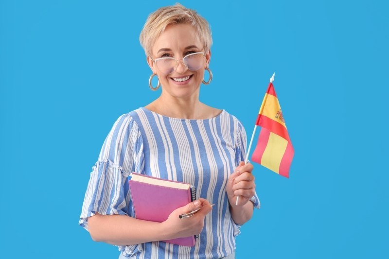 A smiling female teacher stands against a bright blue background, holding a Spanish flag, notebooks, and a pen.