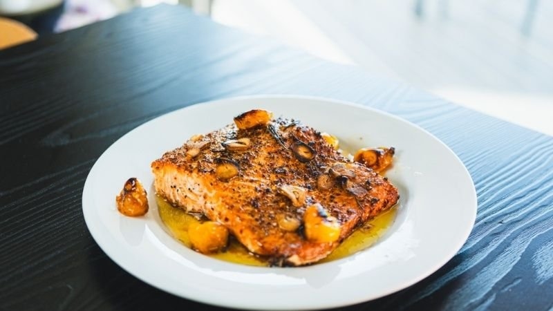 A grilled or roasted salmon fillet served on a white plate, topped with browned ingredients and surrounded by a light sauce or oil.