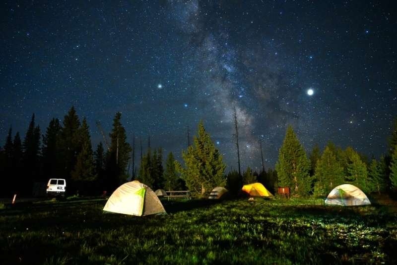 Multiple camping tents set up in a forest clearing under a brilliant star-filled sky and the Milky Way, ideal for stargazing and outdoor adventure.