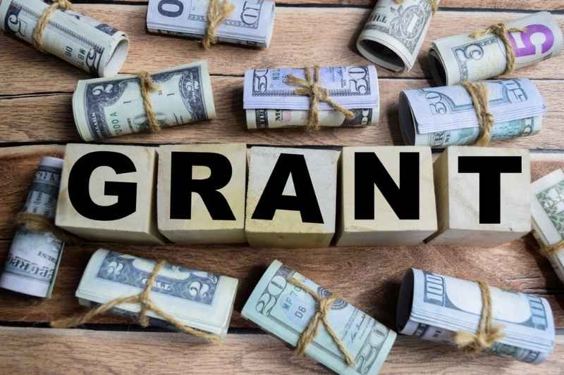 Wooden blocks spelling ‘GRANT’ surrounded by rolled bundles of cash on a wooden table.