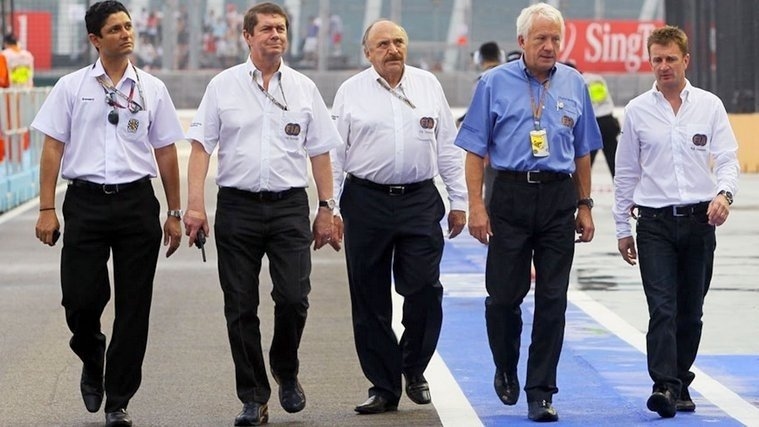 A group of motorsport officials walking together along a Formula 1 circuit, wearing official event uniforms and credentials.
