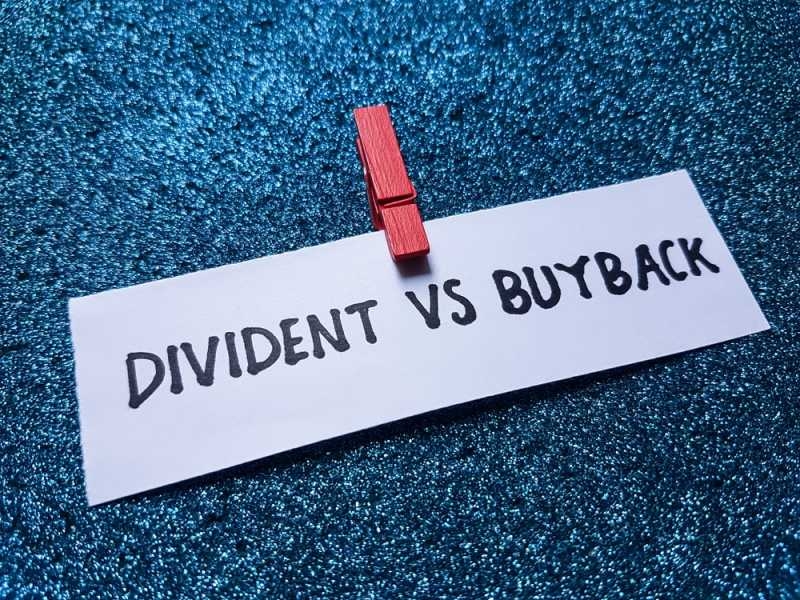 White paper label reading “Dividend vs Buyback” secured with a red clothespin against a textured blue background