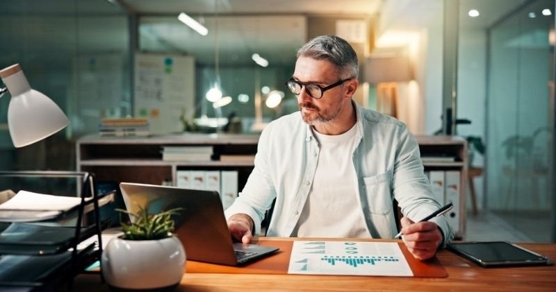 A businessman works at a desk with a laptop, financial charts, and a tablet.