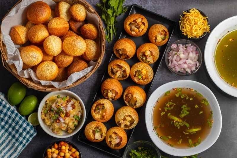Overhead view of assorted Indian street foods, including pani puri, filled puris, chutneys, spiced potatoes, chickpeas, onions, and flavored water.