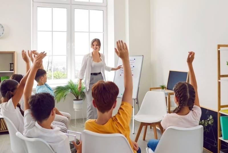 Teacher presenting at the front of a classroom while students raise their hands to answer questions.