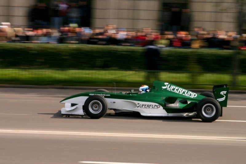 Green and white Formula race car speeding along a city street circuit with motion blur and spectators in the background.