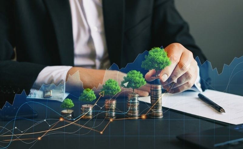 Businessperson stacking coins with small trees to represent sustainable business growth and green investing.