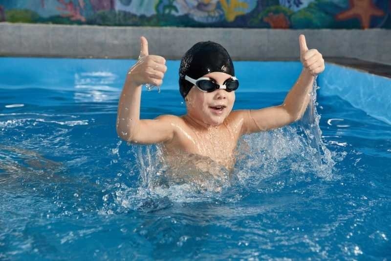 Young swimmer wearing goggles and a swim cap giving two thumbs up while splashing in an indoor swimming pool during a swimming lesson.