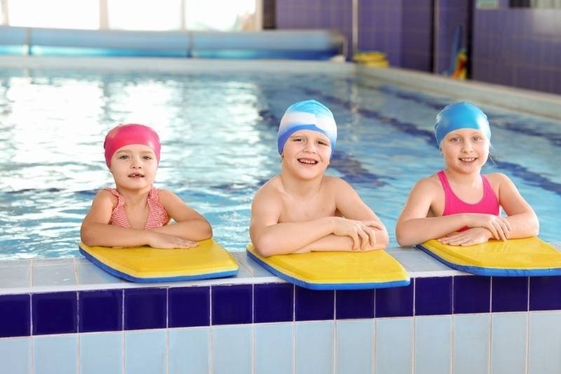 Three children wearing colorful swim caps resting on kickboards at the edge of an indoor swimming pool during a swim lesson.