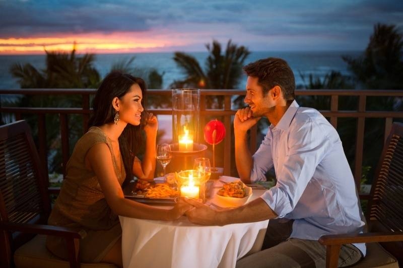 Couple enjoying a romantic candlelit dinner outdoors with an ocean sunset in the background.