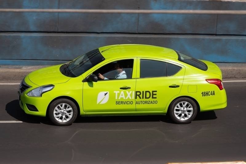 A bright lime-green sedan marked “TAXI RIDE” and “SERVICIO AUTORIZADO” drives along a city road.