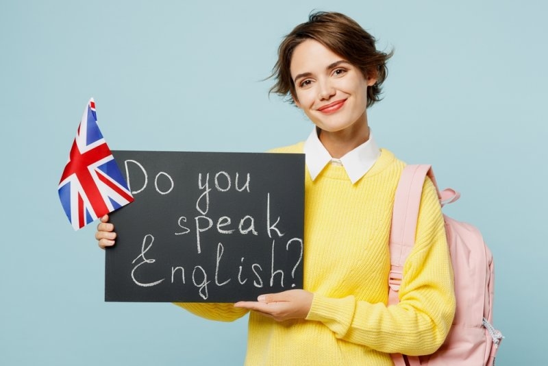 A young woman holding a sign asking, “Do you speak English?” alongside a UK flag.