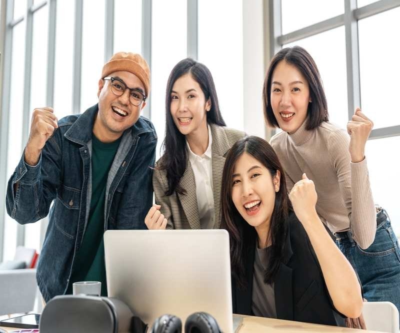 A diverse group of coworkers smiling and celebrating together around a laptop in an office.