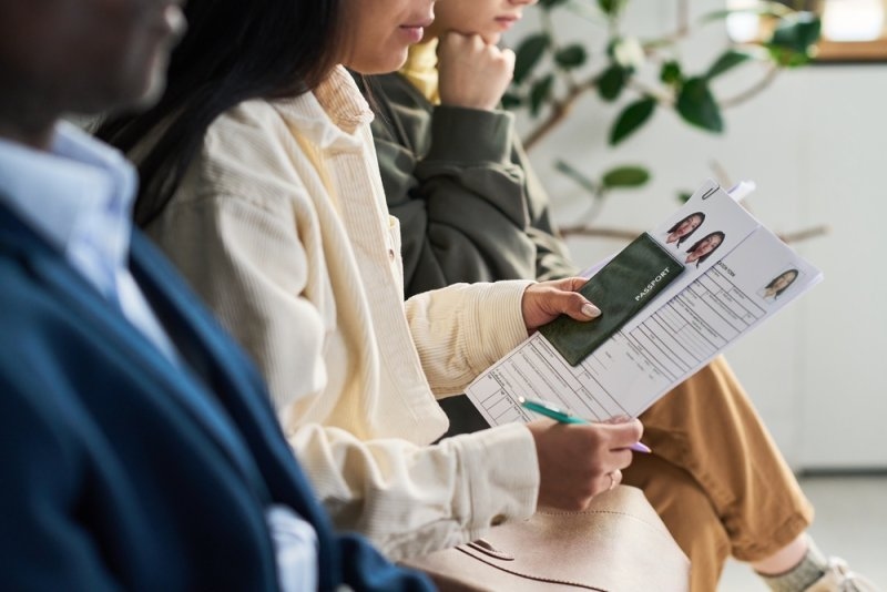 A young group of girls sitting in visa center waiting area, and a woman holding passport and visa application form with pen, preparing documents for processing.