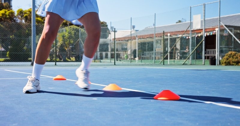Tennis player practicing footwork drills around colorful training cones on an outdoor tennis court.