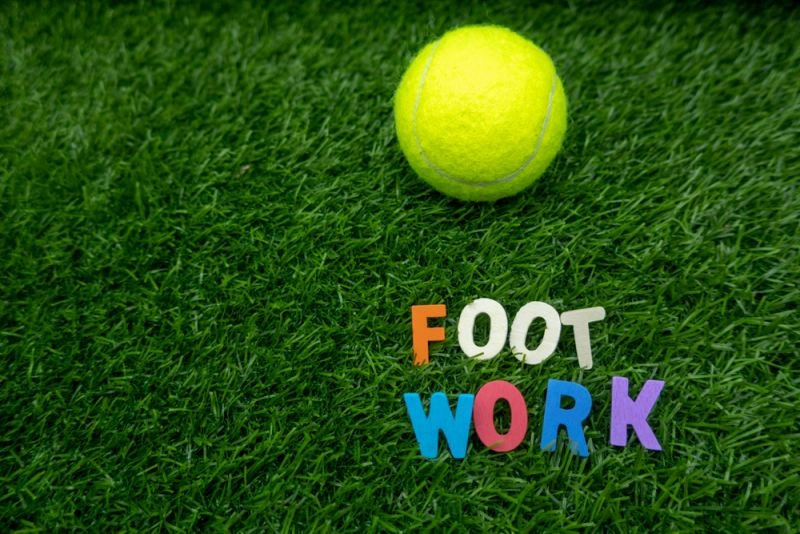 Yellow tennis ball resting on grass beside colorful letters spelling “FOOT WORK.”