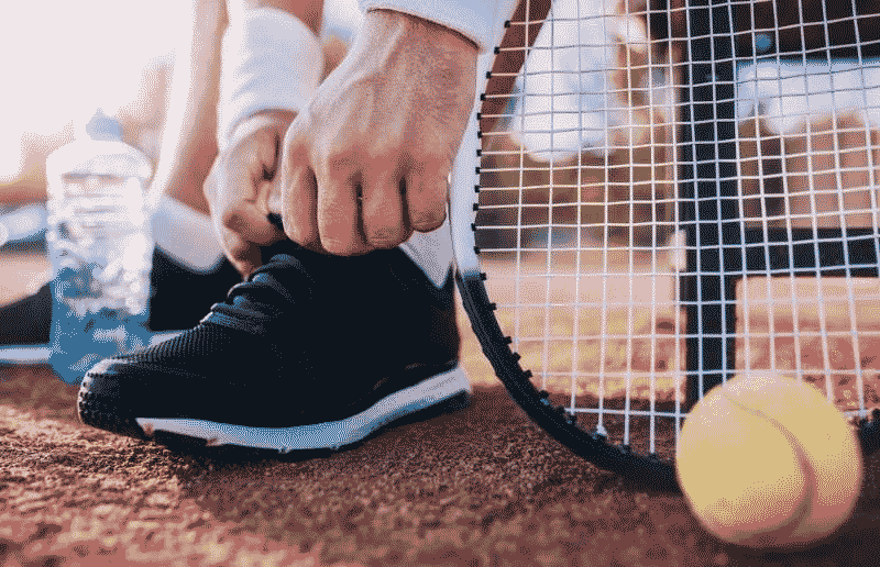 Close-up of a tennis player tying black athletic shoes on a tennis court, with a racket and tennis ball nearby.