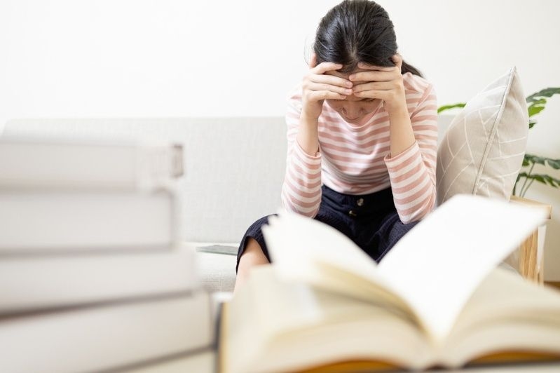 Young woman sitting with head in hands, looking overwhelmed while studying near books.