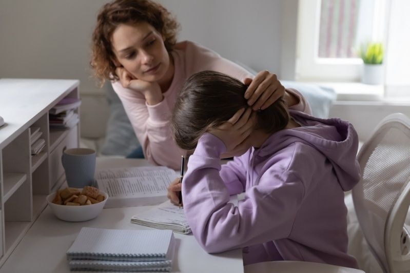 Child looking stressed while studying at a desk, with an adult offering support.