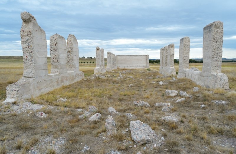 Weathered concrete ruins and stone structures standing in an open grassy field beneath a cloudy sky.