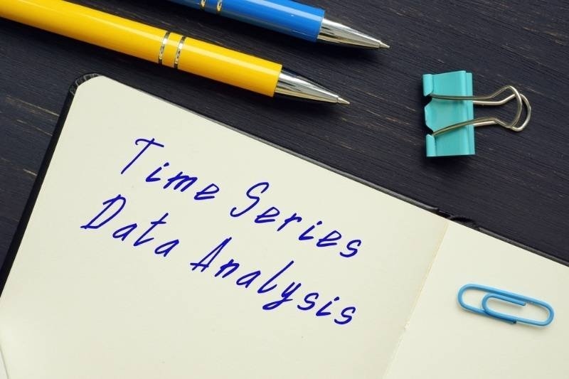 Open notebook displaying the handwritten title "Time Series Data Analysis" surrounded by pens, a binder clip, and a paper clip on a desk, symbolizing data analytics and statistical modeling.