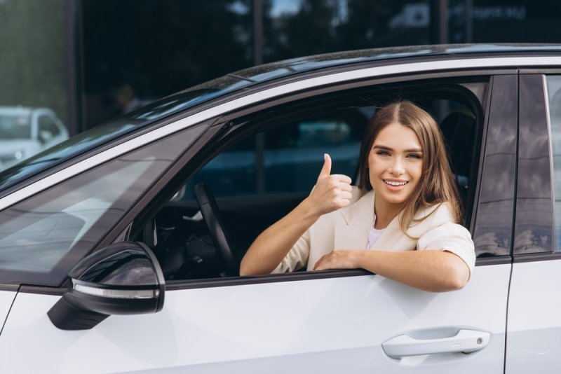 A smiling woman sits in the driver's seat of a white SUV and gives a thumbs-up through the open window.