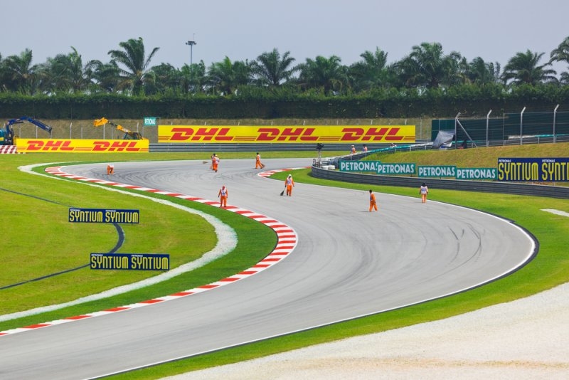 Formula 1 race circuit with sweeping corners, red-and-white curbs, safety barriers, palm trees, and track marshals.