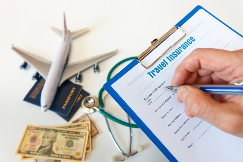 Person filling out a travel insurance application on a clipboard beside an airplane model, passports, a stethoscope, and US dollar bills.