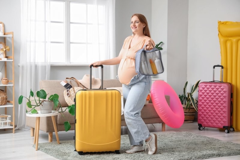 Pregnant woman preparing for travel at home with luggage, a travel bag, and an inflatable neck pillow.
