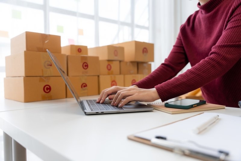 Person working on a laptop at a desk with stacks of cardboard shipping boxes in the background.