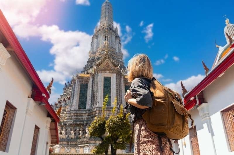 Traveler with a backpack looking up at the ornate architecture of Wat Arun temple on a bright sunny day in Bangkok.